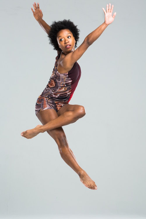 A female dancer jumping in mid-air. Her torso twists away from the camera while her left calf rests on top her right thigh. Her arms are out-stretched.